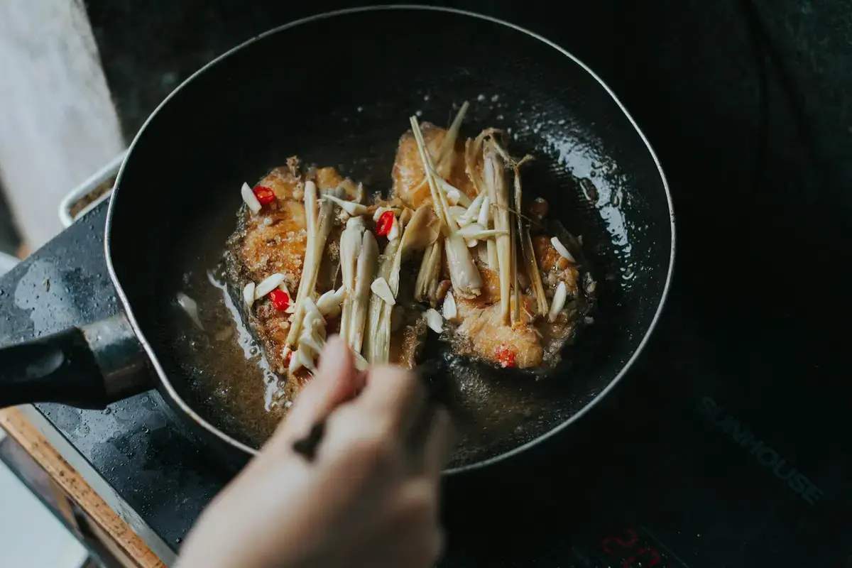 熱鍋中以少量油爆香蒜片、辣椒與香茅，一旁是煎得金黃的魚排（示意圖）