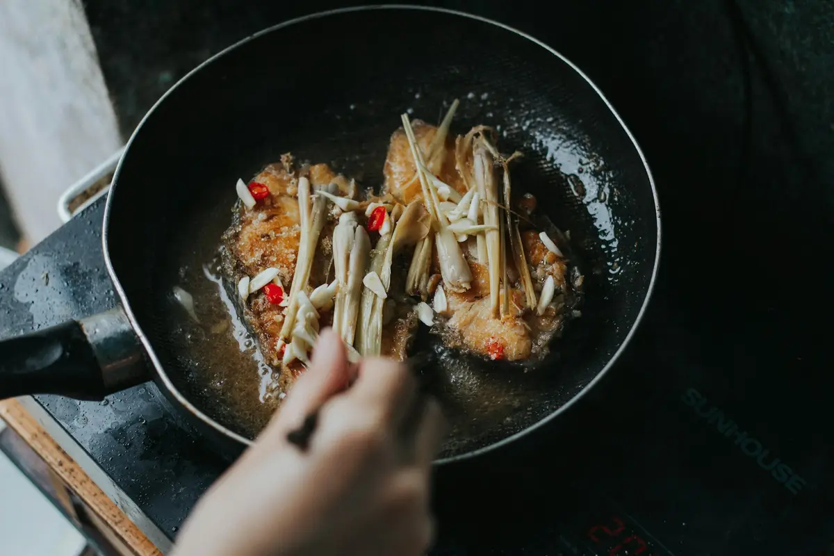熱鍋中以少量油爆香蒜片、辣椒與香茅，一旁是煎得金黃的魚排（示意圖）