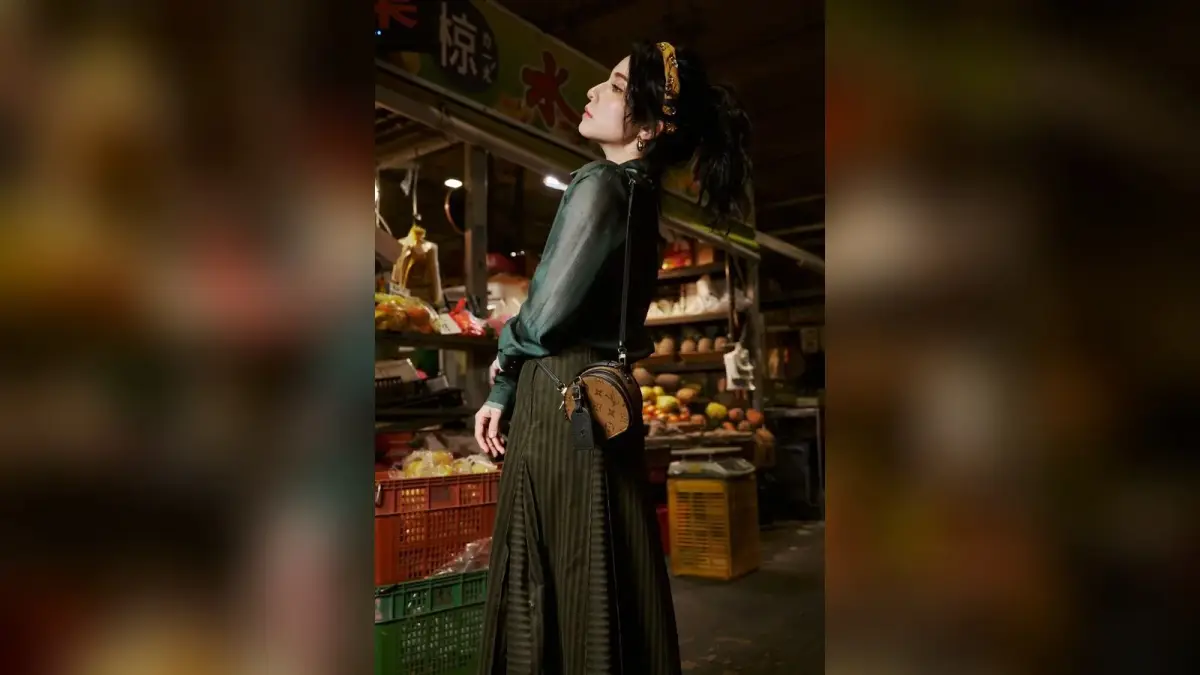 穿深綠色衣服、配戴髮飾與肩背包的人物站在蔬果市場，光線照亮側臉與衣料。