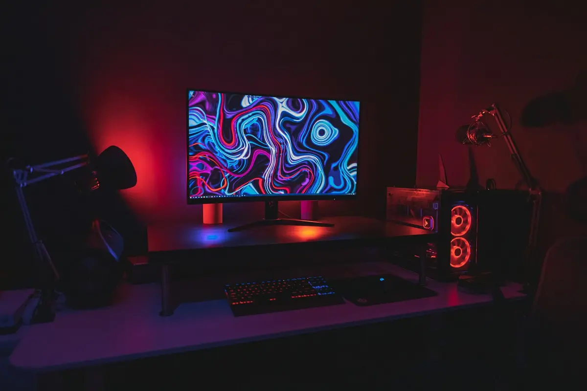 昏暗的遊戲工作桌上，電腦螢幕顯示抽象彩色圖像，旁有鍵盤與桌上型電腦。