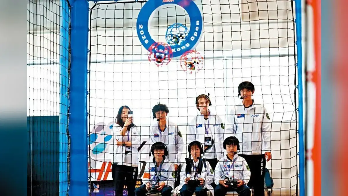 四名台灣青少年選手與教練在香港無人機足球賽場手持獎盃，前方放著球形防護籠無人機。