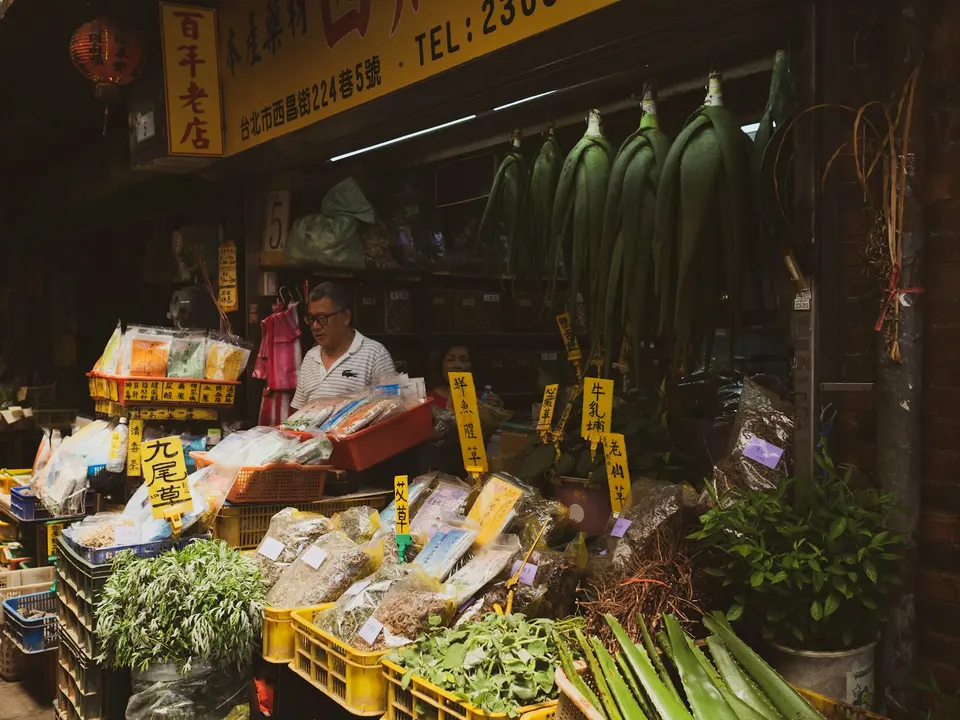 台北中藥材老店實景,男性店主站在擺滿各式乾燥藥材的攤位前,掛有中文招牌