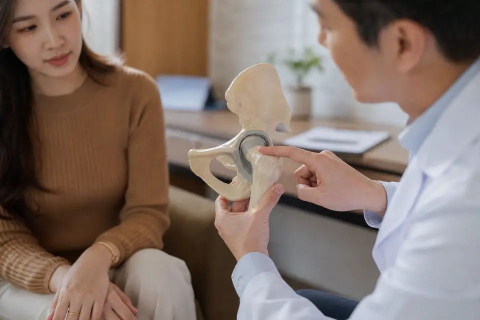 醫師在診間拿著髖關節骨骼模型，向坐著的患者說明疼痛位置與就醫判斷（示意圖）