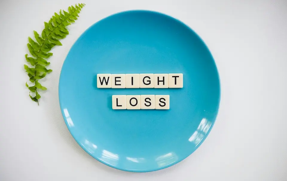 淺藍色圓形餐盤放在白色背景上,盤中用白色方形字母磚拼出「WEIGHT LOSS」字樣,旁邊點綴綠色植物葉片