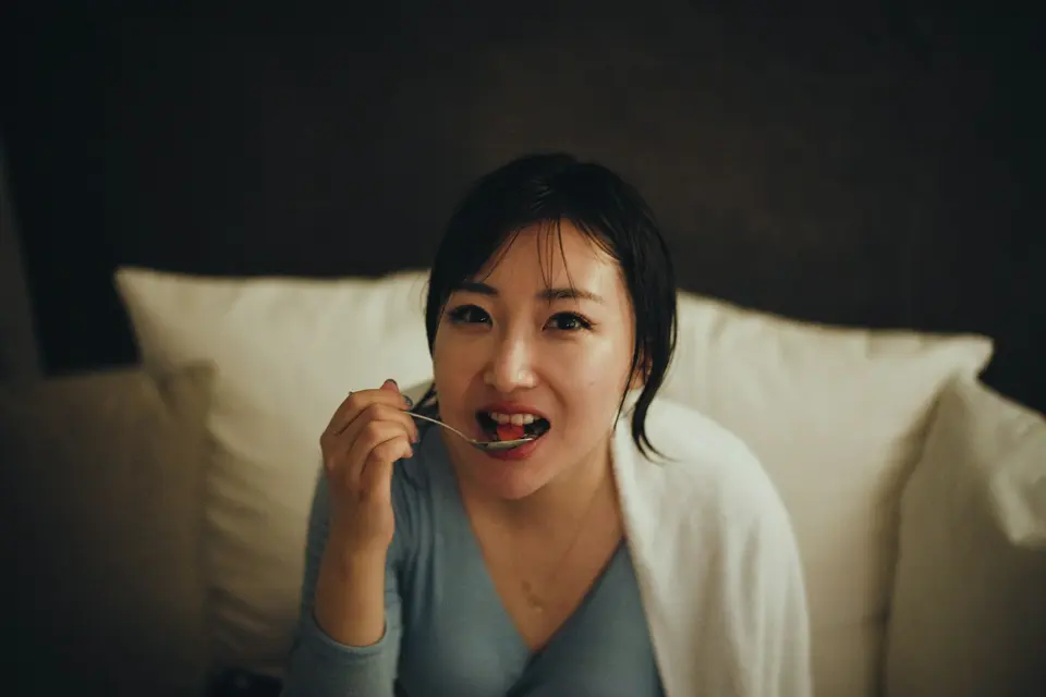 一位女性坐在床上,神情略顯疲憊,手持湯匙,身披白色毯子