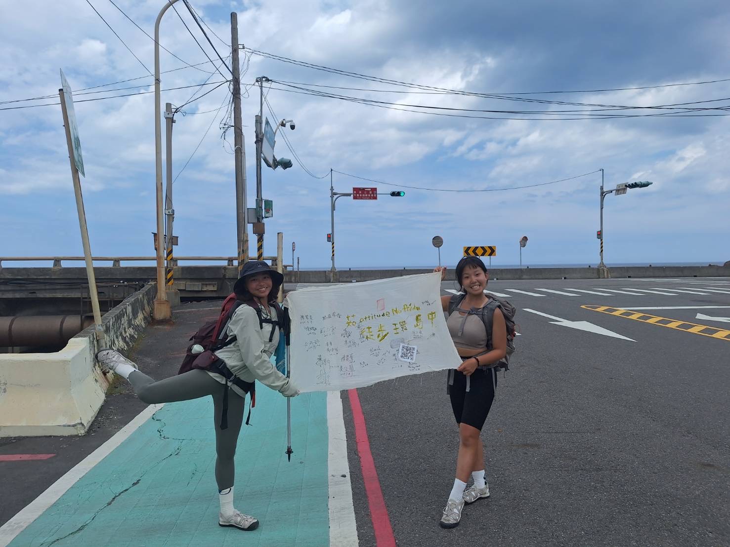 兩位徒步者在海邊道路展示旅程布旗，背景是海岸公路與天空