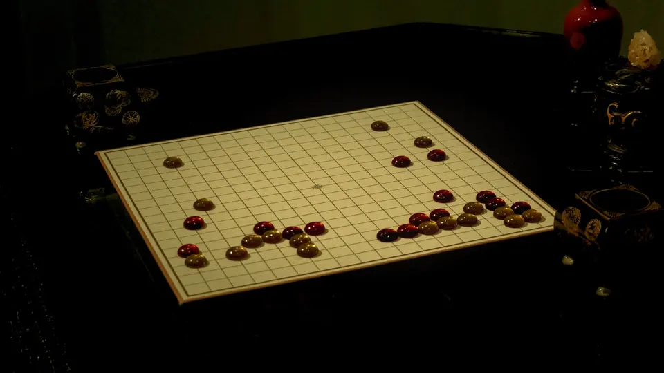 暗室木桌上擺放著白色格線圍棋棋盤,棋盤上散落著紅褐色棋子,兩側各有一個精緻黑色棋盒