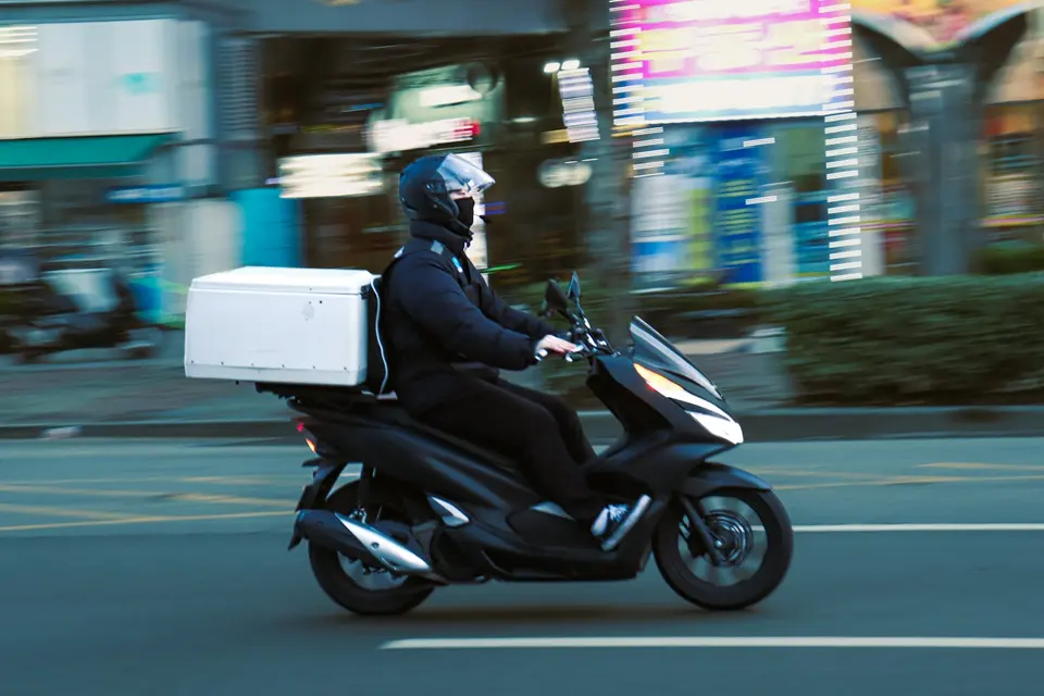 外送員騎黑色機車在城市街道疾馳,背後背著大型白色保溫外送箱