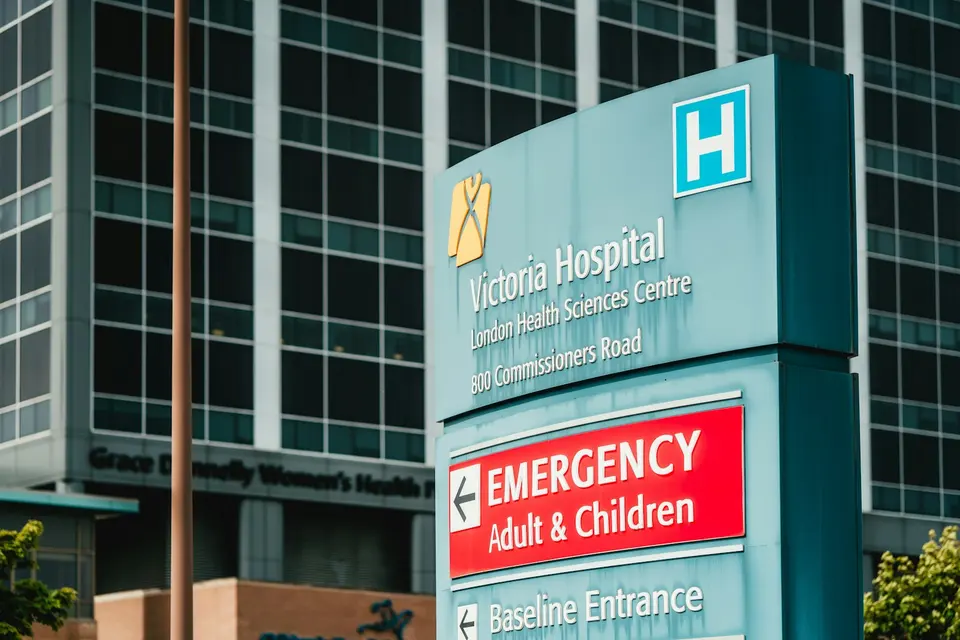 醫院門口的急診指示牌,藍色立柱上標示 Victoria Hospital 與紅底白字的 EMERGENCY Adult & Children 方向箭頭,背景為現代化玻璃帷幕大樓