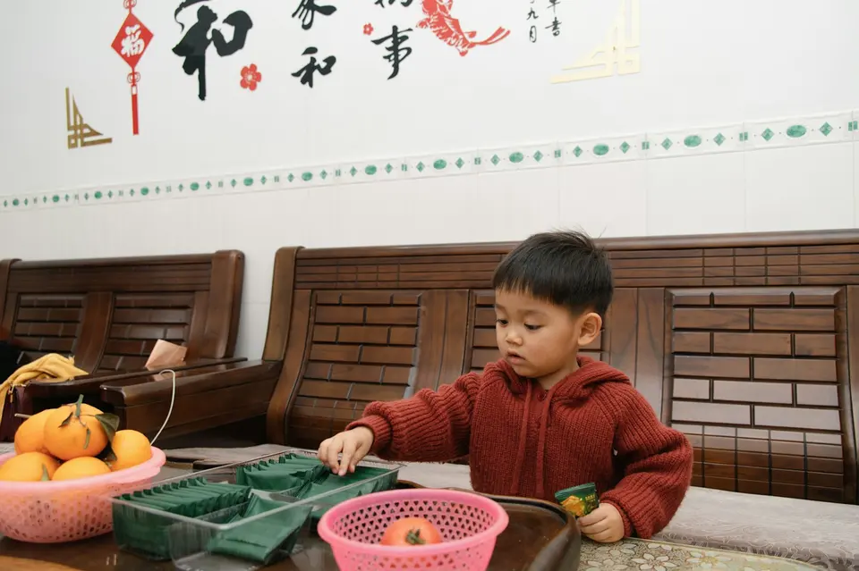 一個穿紅色針織外套的亞洲小男孩坐在木桌前,專注地撥弄桌上的分格盒子,旁邊擺著橘子,牆上有中文書法裝飾